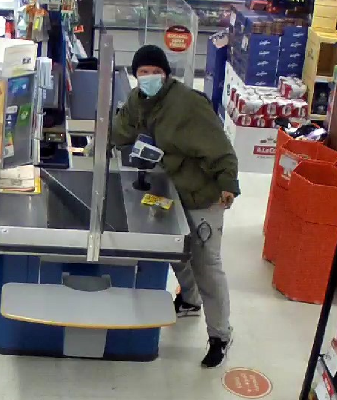 Mies kaupan kassalla vihreässä takissa ja harmaissa housuissa ottamassa jotain kassasta.