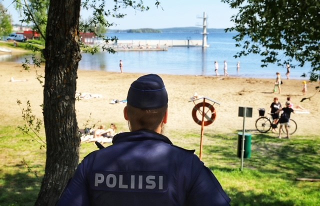 Poliisi valvoo järjestystä kesäisellä uimarannalla. Poliisi seisoo selin kameraan suikka päässä, haalareissa lukee selässä poliisi.