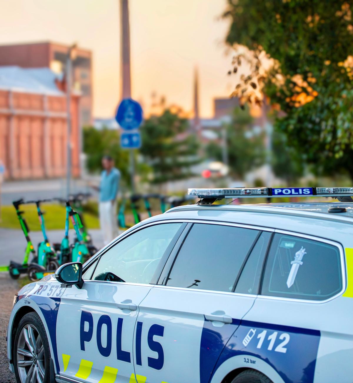 Poliisiauto etualalla, taustalla useita sähköpotkulautoja ja ihminen, joka seisoo kadulla.