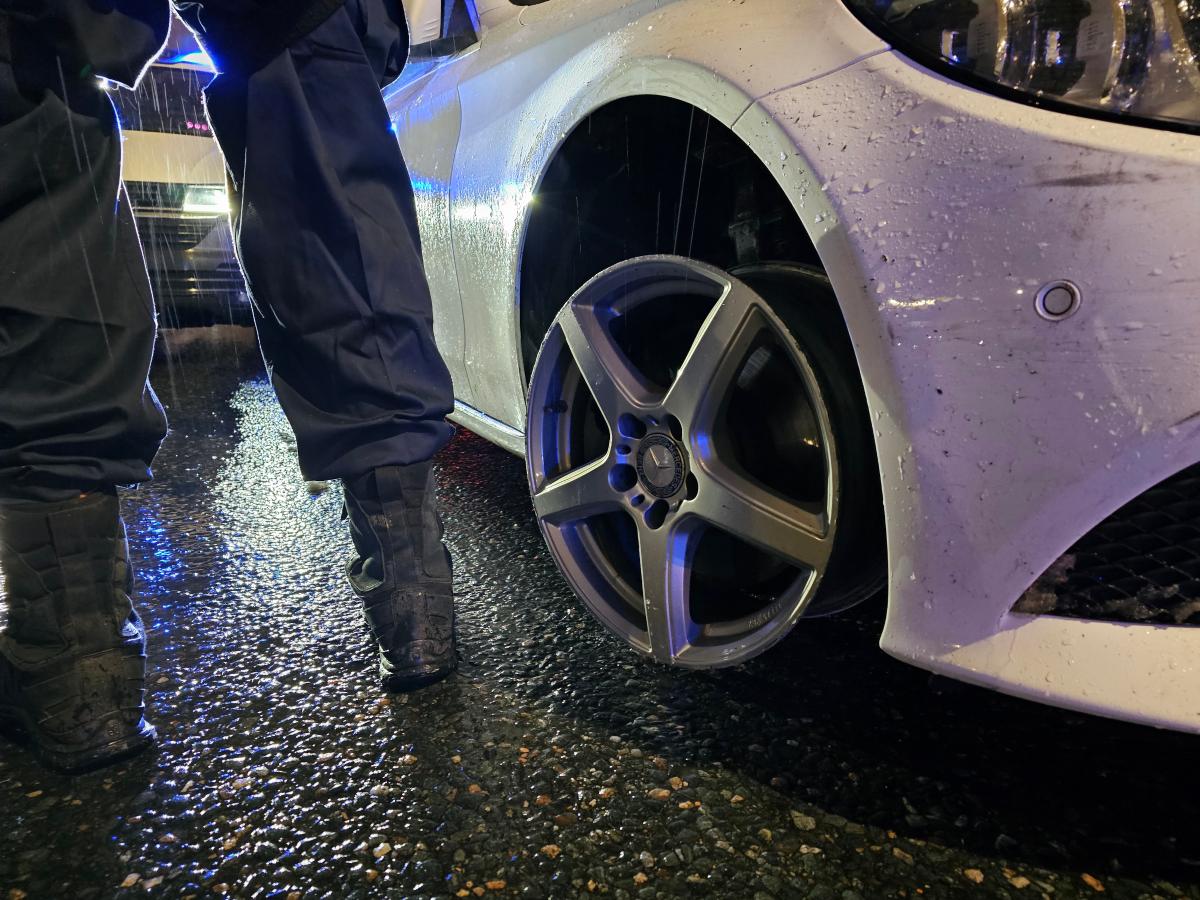 Anastetun ajoneuvon oikea eturengas, poliisimiehen jalat, takana poliisiauton hälytysvalot