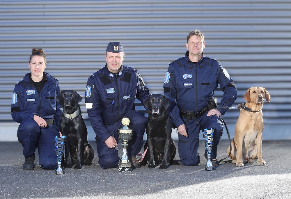 Kolme virkapukuista poliisia on kyykistyneinä, jokaisella on vierellään poliisikoira.