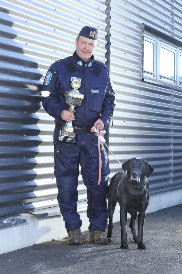 Virkapukuinen poliisi ja poliisikoira seisovat seinustalla. Poliisilla on pokaali kädessään.