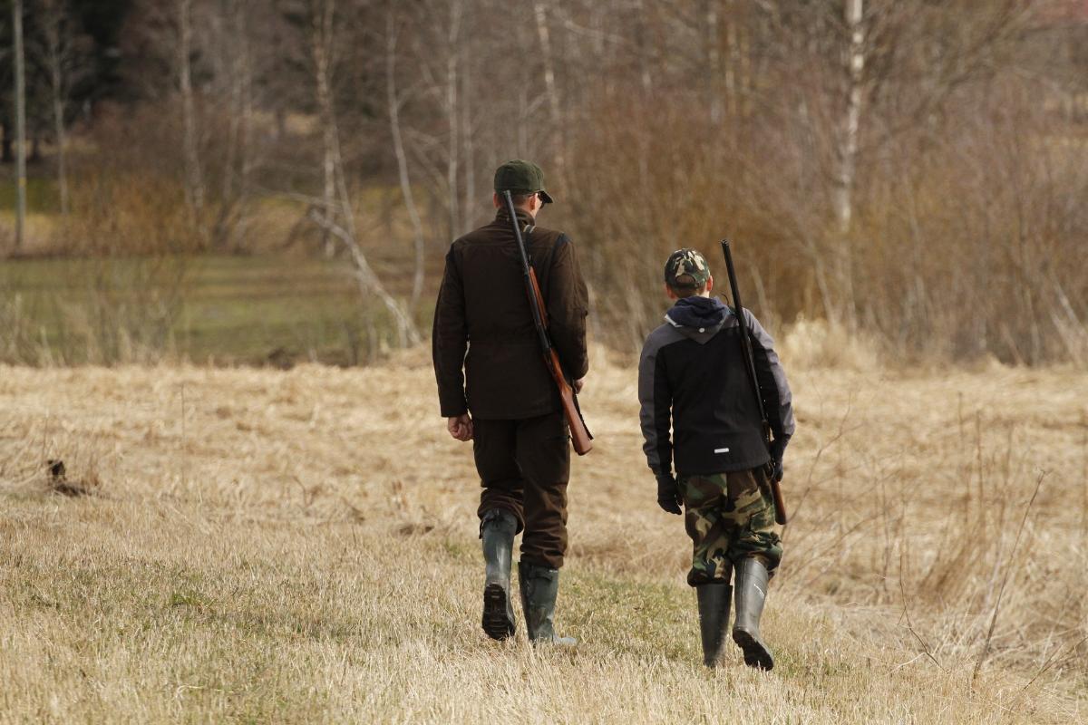 Mies ja poika kävelevät kellastuneella pellolla aseet selässään.