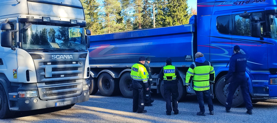 Poliisin ja Tullin työntekijöitä valvomassa raskasta liikennettä. Etualalla valkoinen rekka ja taka-alalla tummansininen kuorma-auto.