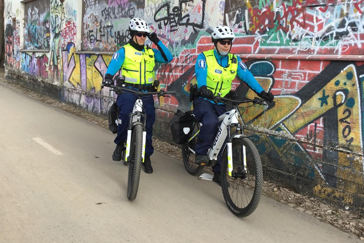 Pyörätiellä rakennuksen vieressä on kaksi poliisia polkupyörän selässä huomioliivit yllään ja kypärät päässään.