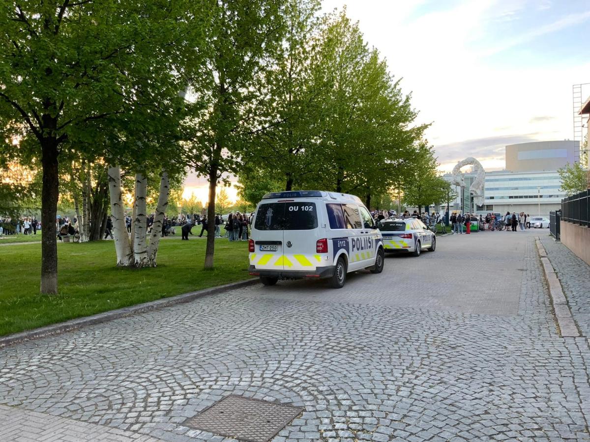 Kaksi poliisiautoa kadun varressa puiston vieressä. Taustalla on paljon nuoria ihmisiä.