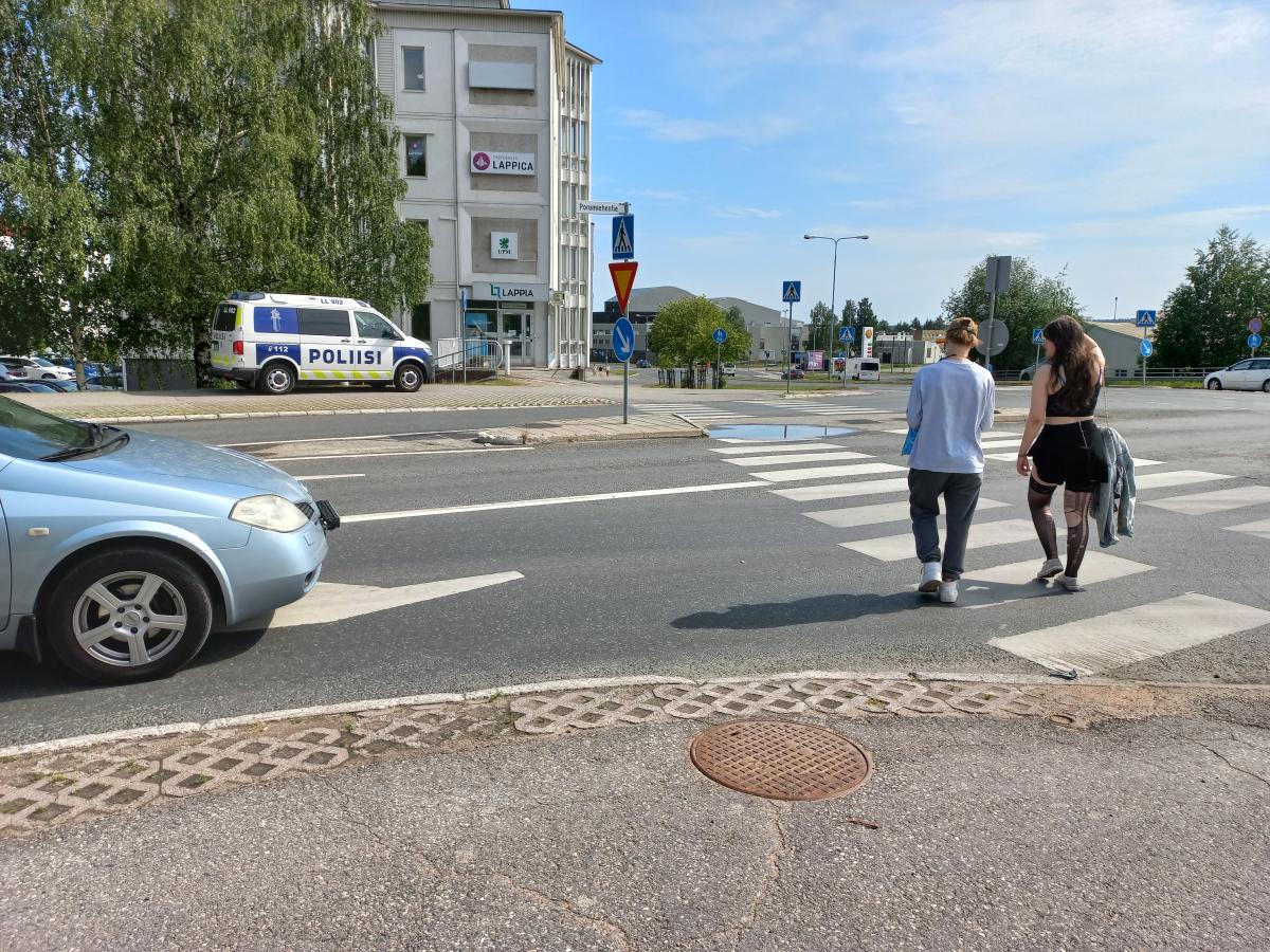Kaksi ihmistä kävelee suojatien yli, kun auto seisoo suojatien edessä. Poliisiauto seisoo suojatien toisessa päässä kadun varressa.