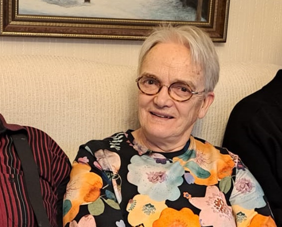 Harmaahiuksisella naisella on yllään kukallinen pusero ja päässään pyöreäkehyksiset silmälasit.