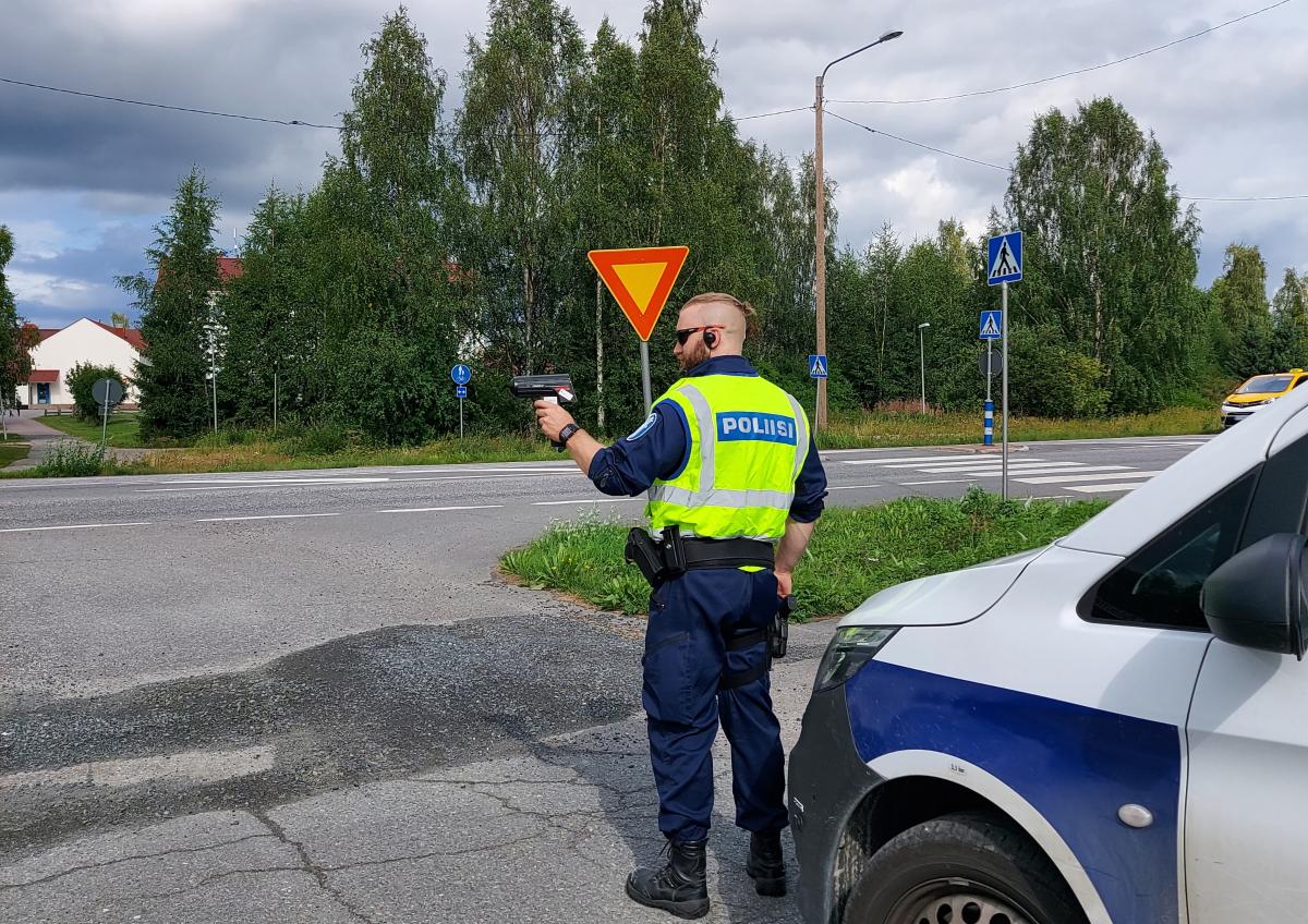 Poliisimiehellä on kädessä tutka ja hän seisoo tien laidassa. Takana on poliisiauto.