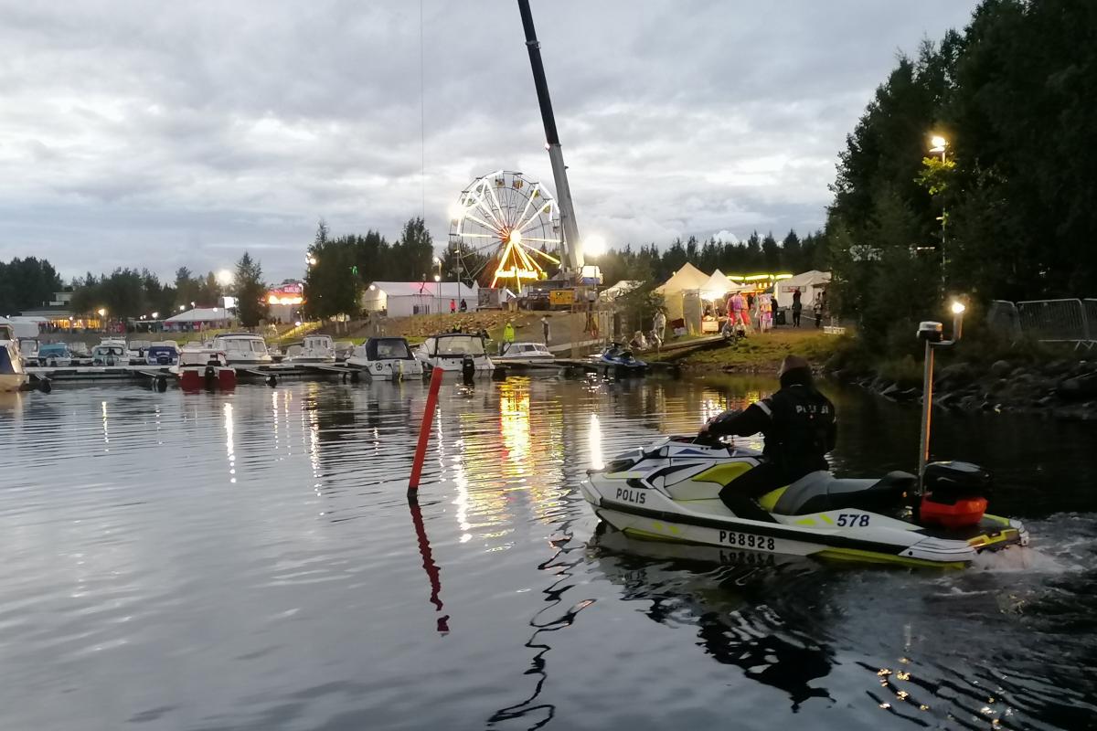 Poliisi istuu vesijetin kyydissä rannan tuntumassa. Laiturissa on useita veneitä. Rannalla on maailmanpyörä ja festivaalitelttoja.