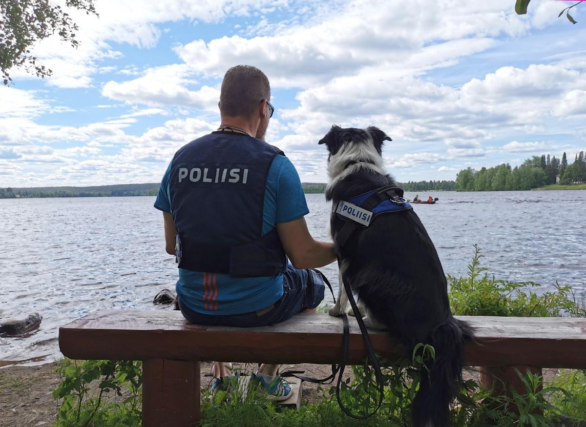 Poliisi ja poliisikoira istuvat penkillä joen rannassa ja katsovat joella kulkevaa venettä.