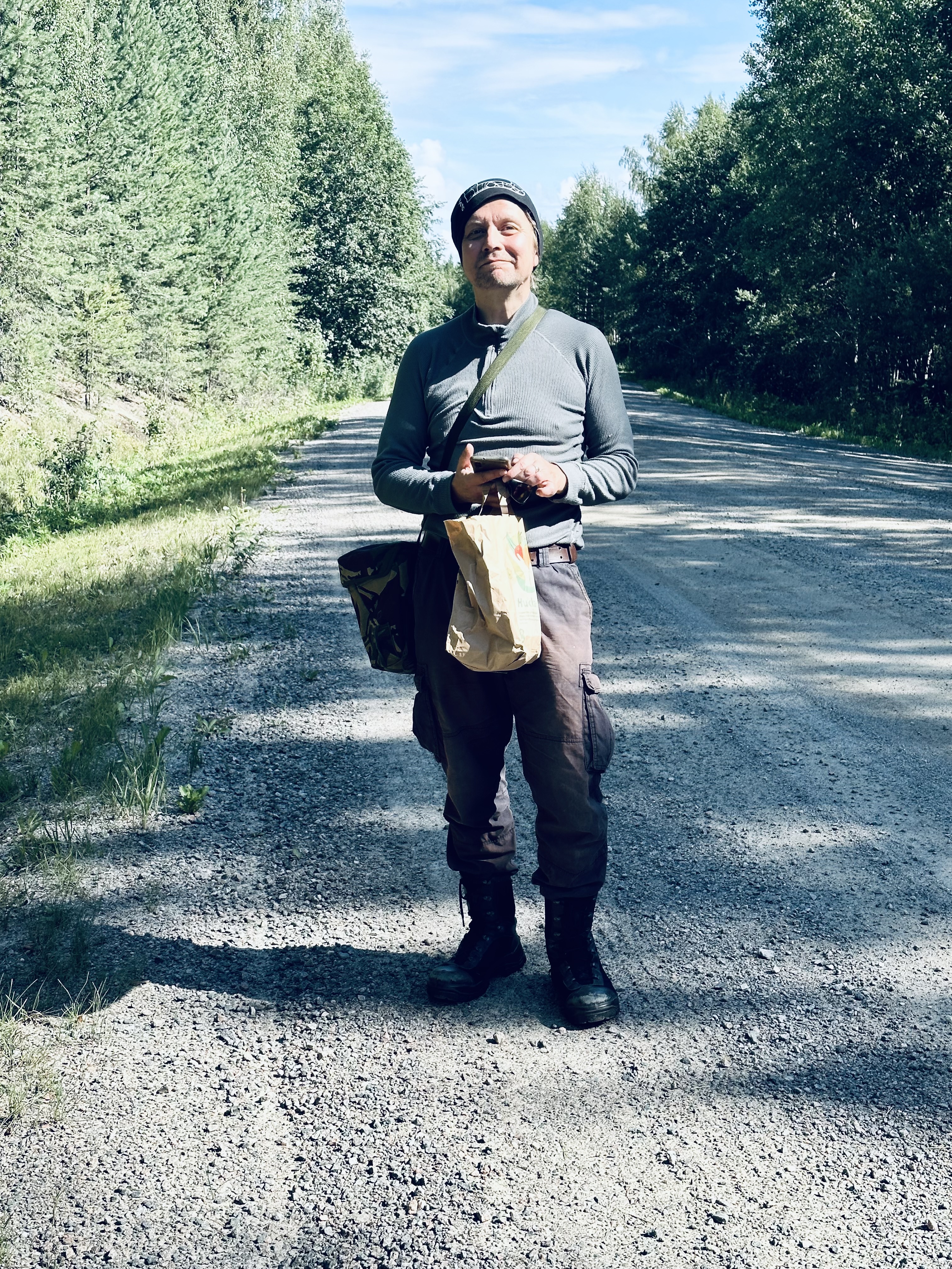 Henkilö seisoo hiekkatiellä. Tien sivuilla on havupuita. Yllä hänellä on tumma pipo, tumma korkeakauluksinen paita, reisitaskuhousut ja maihinnousukengät. Oikean reiden vieressä on olkalaukku, jonka hihna on vasemmalla olalla. Kädessä hänellä on paperipussi. Hän pitelee käsissään myös älypuhelinta.