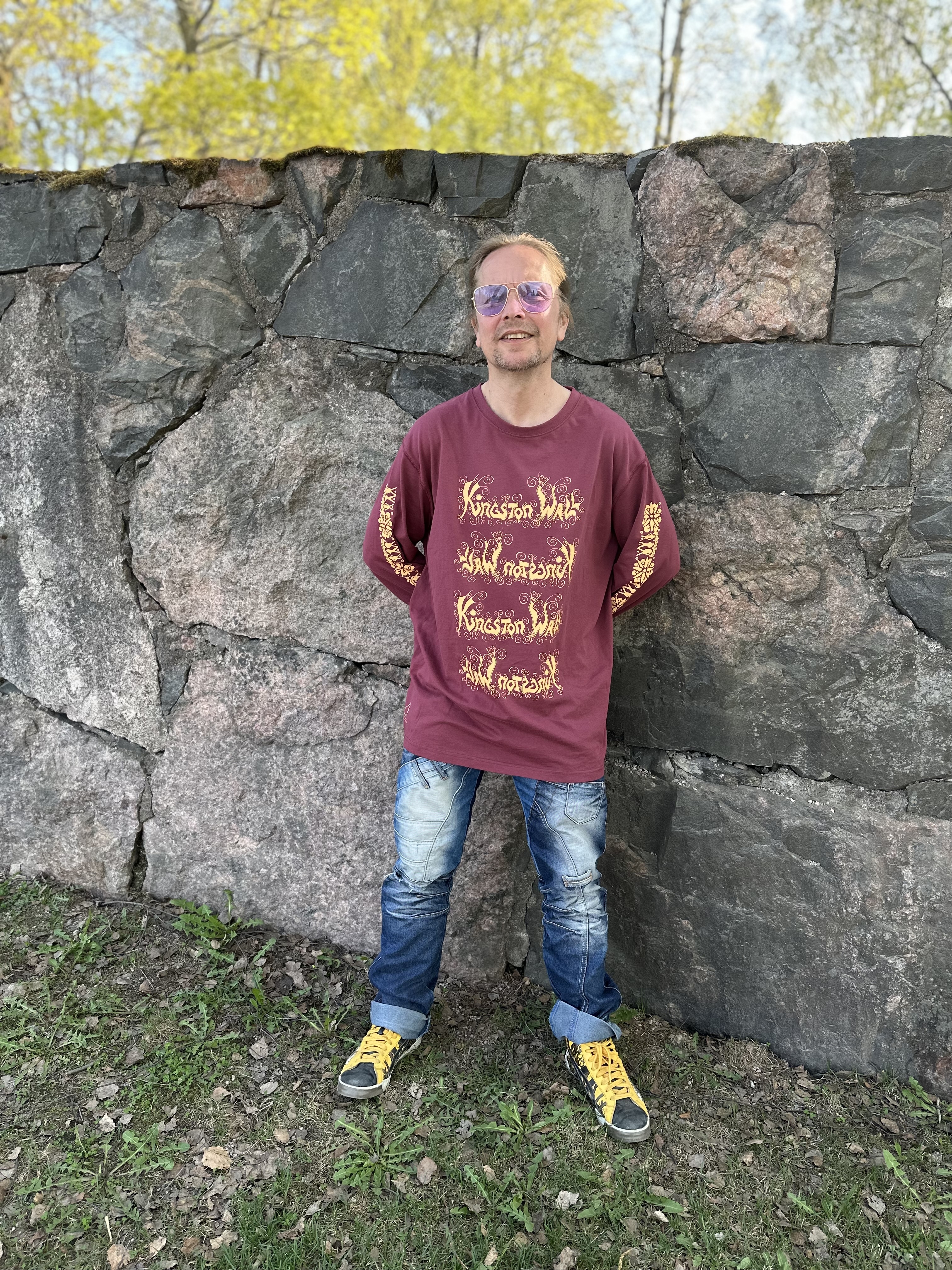 Henkilö seisoo korkeaa kivimuuria vasten. Muurin takana näkyy lehtipuita. Maassa on ruohoa ja multaa. Yllä henkilöllä on aurinkolasit, viininpunainen pitkähihainen, jossa lukee keltaisella Kingston Wall neljä kertaa edessä niin, että joka toinen teksti on väärin päin. Hihoissa on keltaista kuviota. Jalassa on siniset, reisistä kulutetut farkut. Kengät ovat tummat ja niissä on keltaiset nauhat. Kädet ovat selän takana, ja henkilö hymyilee niin, että hampaat näkyvät.