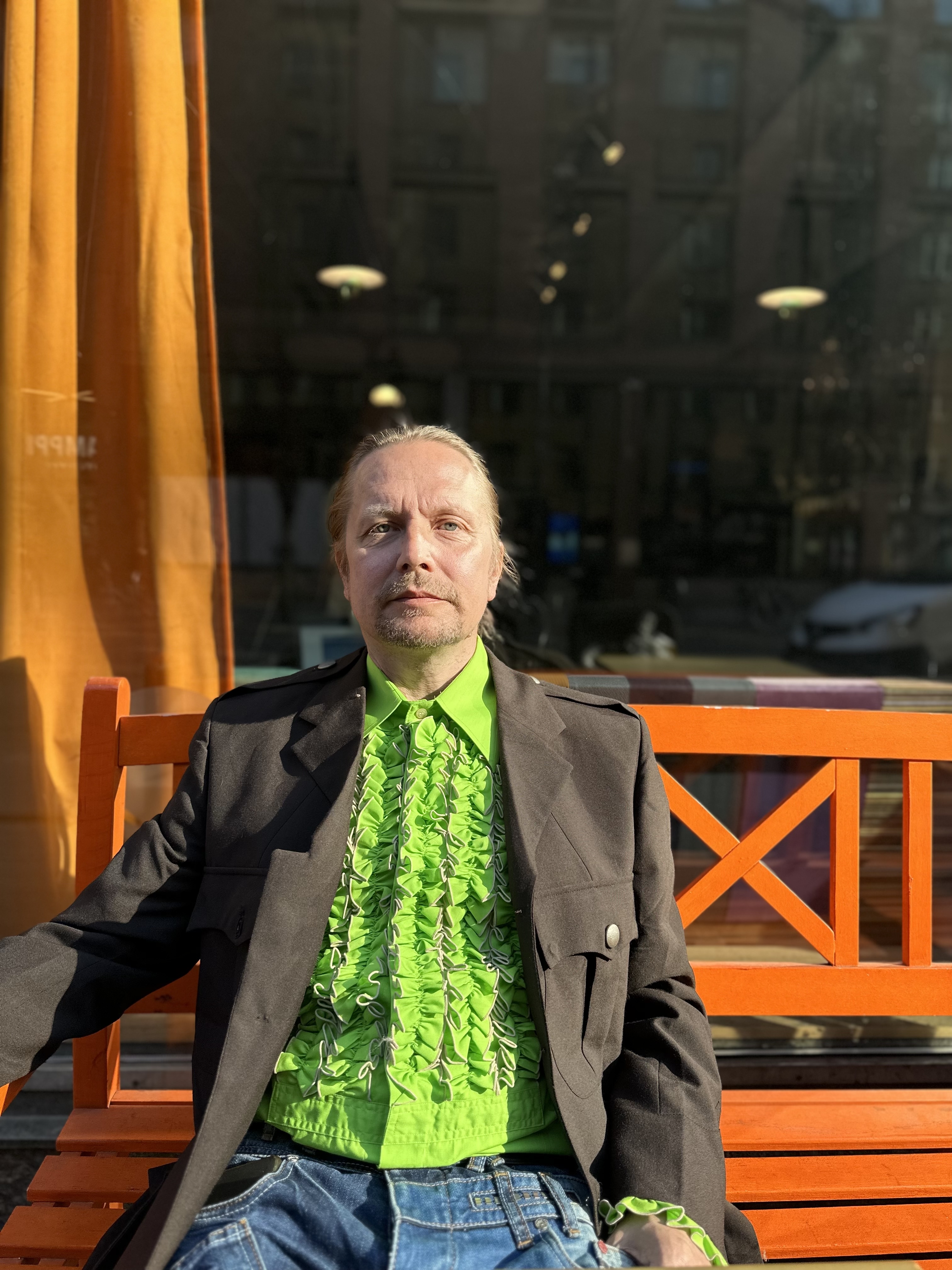 Henkilö istuu punertavalla puupenkillä ulkona. Takana on suuri ikkuna, jossa on oranssi verho sivussa. Ikkunasta heijastuu kerrostalo ja valoja. Henkilöllä on vaalea tukka ja lyhyt vaalea parta. Yllä hänellä on limenvihreä röyhelöinen paita, tumma takki ja siniset farkut.