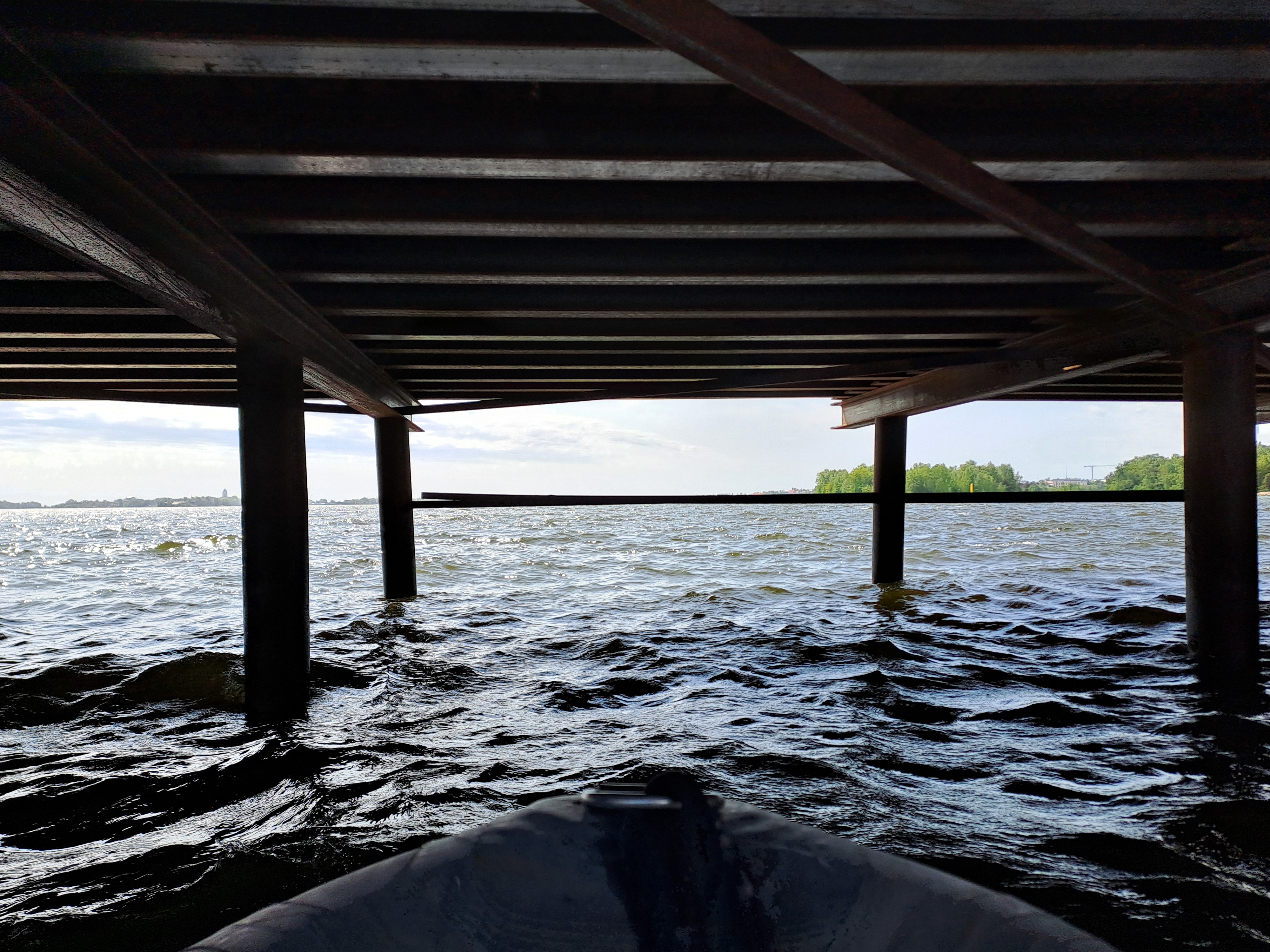 Siltarakenteiden alla näkyy veneen nokka vedessä. Veneen edessä jonkin matkan päässä on vaakatasossa oleva palkki. Taustalla näkyy taivasta ja oikealla puita.