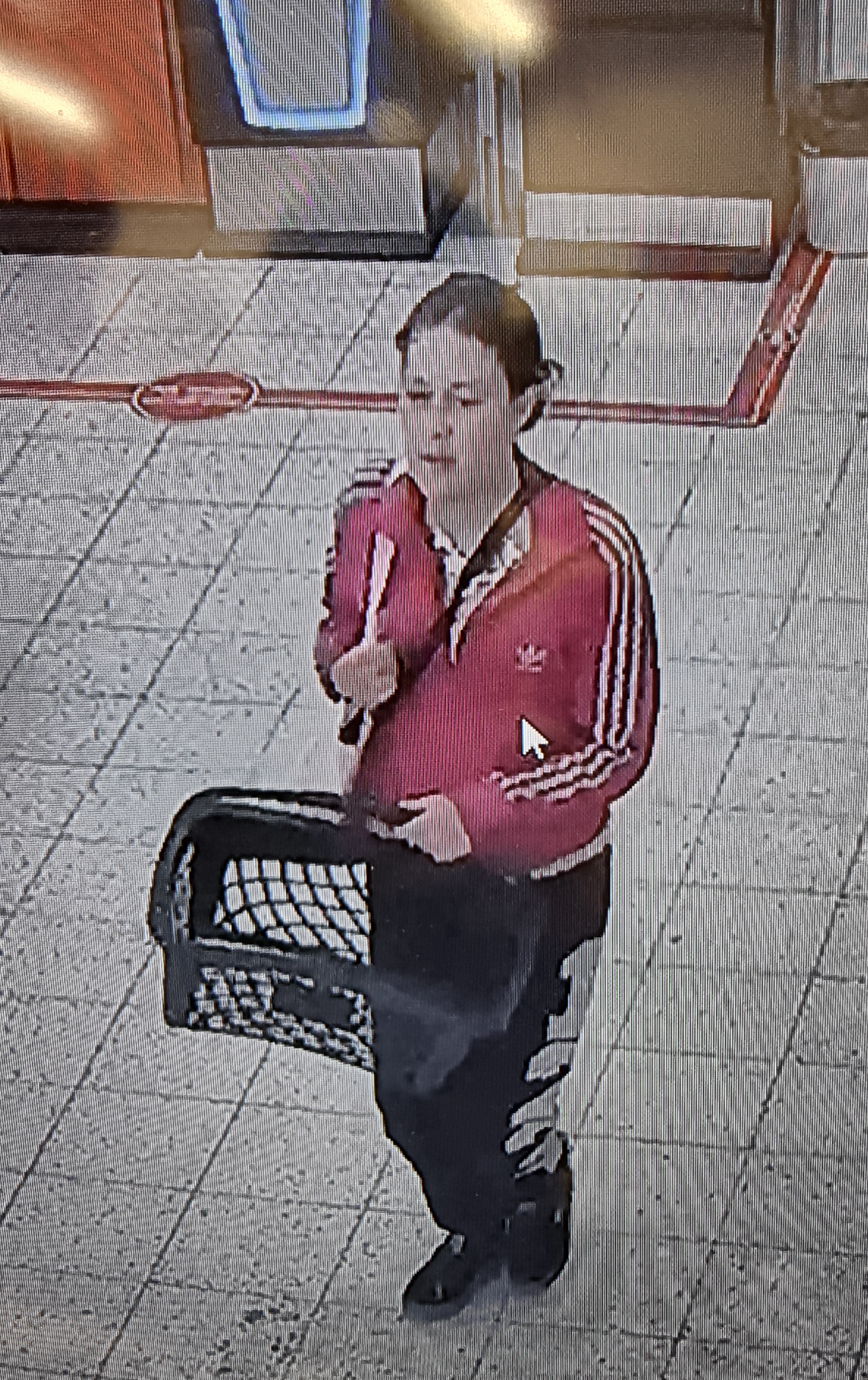 Nainen punaisessa pusakassa kävelee ostoskorin kanssa. Hiukset ovat kiinni. Jalassa mustat housut.