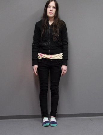 Nainen seisoo tummissa vaatteissa harmaata seinää vasten.