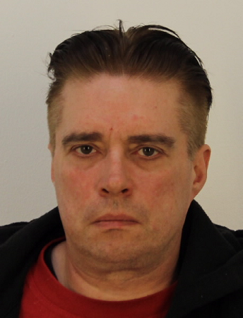 Tummahiuksinen mies katsoo kameraan. Kuvassa näkyvät kasvot ja hieman punaista ja mustaa paitaa.