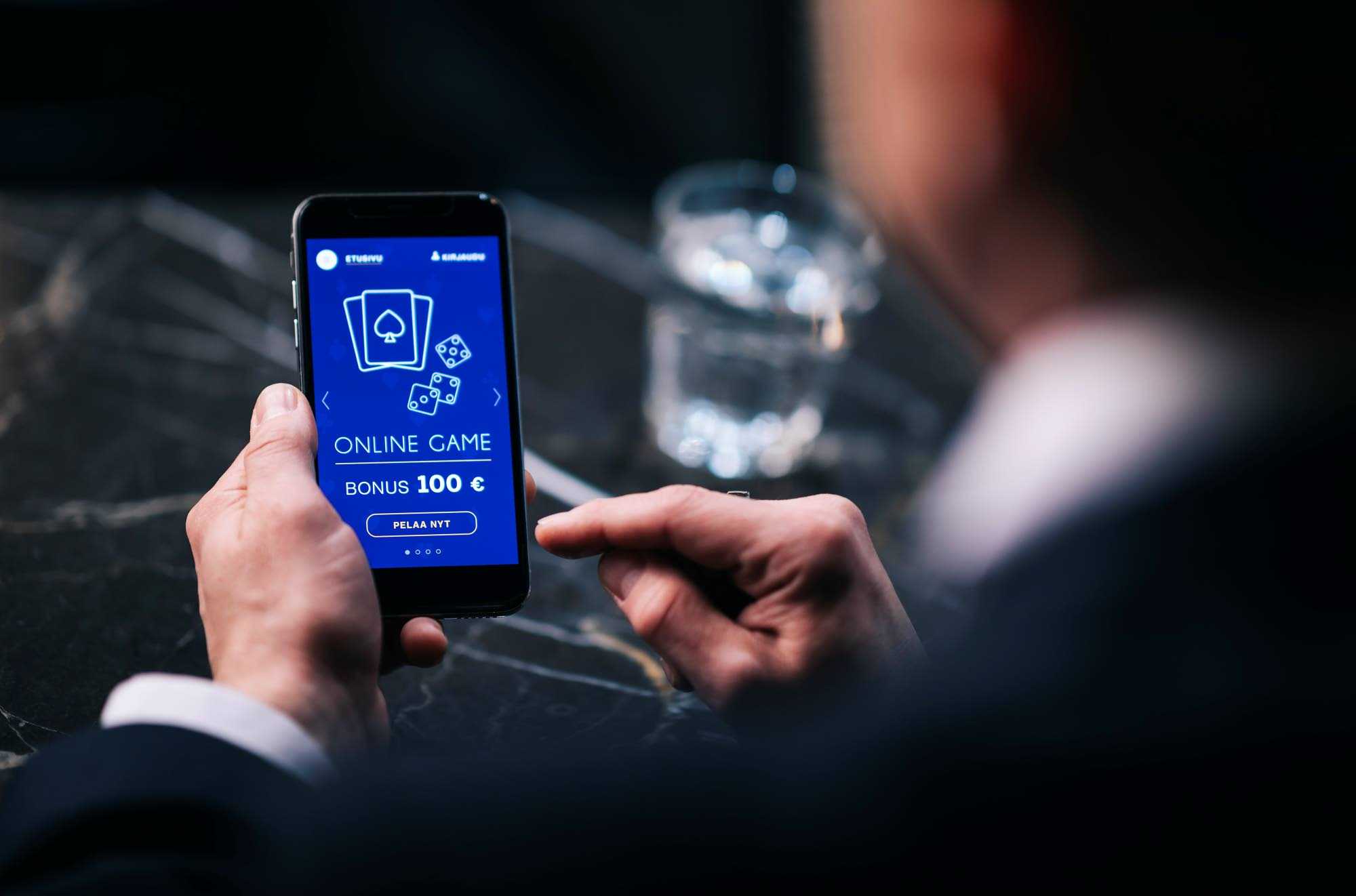 En person håller en mobiltelefon i handen, på telefonskärmen finns penningspelsajtens text Online Game Bonus 100 € Pelaa nyt (Spela nu).