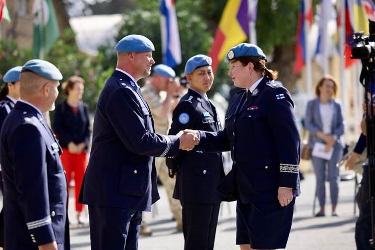 Finnish police officer Satu Koivu shaking hands. In the picture, everyone is wearing blue feathers and uniforms. In the background are flags of different countries and photographers.